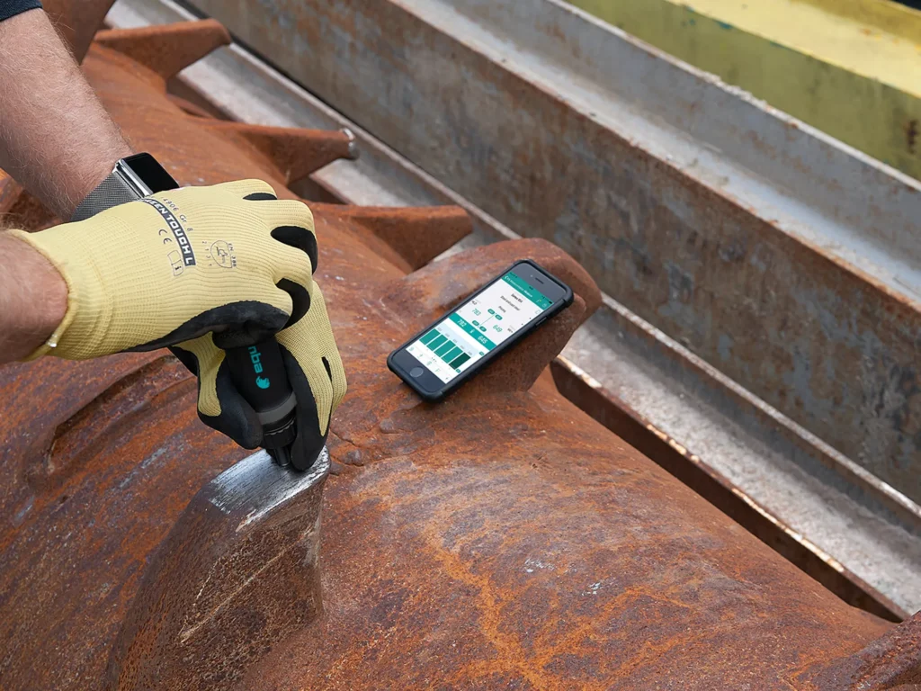 Un técnico con guantes realiza una prueba de dureza portátil en una gran pieza metálica oxidada. Un dispositivo inteligente muestra los resultados de la medición.