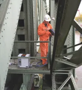 Técnico aplicando espectrometría sobre una estructura metálica de puente para analizar la composición de materiales en exteriores
