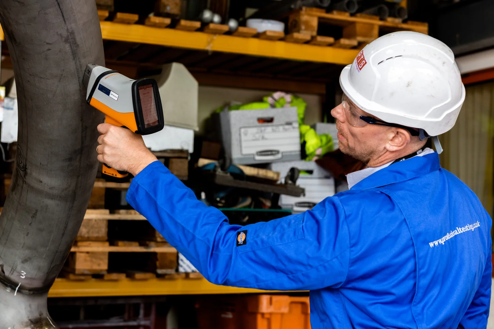 Un técnico con equipo de seguridad realiza un análisis de fluorescencia de rayos X en una tubería metálica dentro de un almacén industrial.