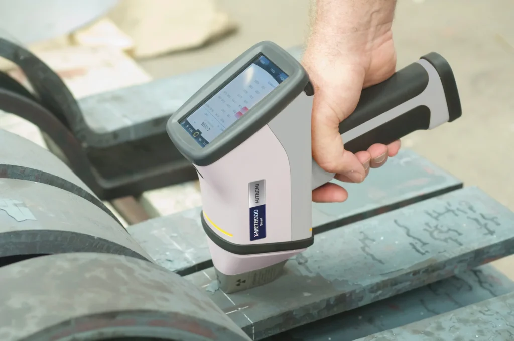 Una mano sostiene un analizador portátil XRF de Hitachi para el análisis de fluorescencia de rayos X sobre piezas metálicas.