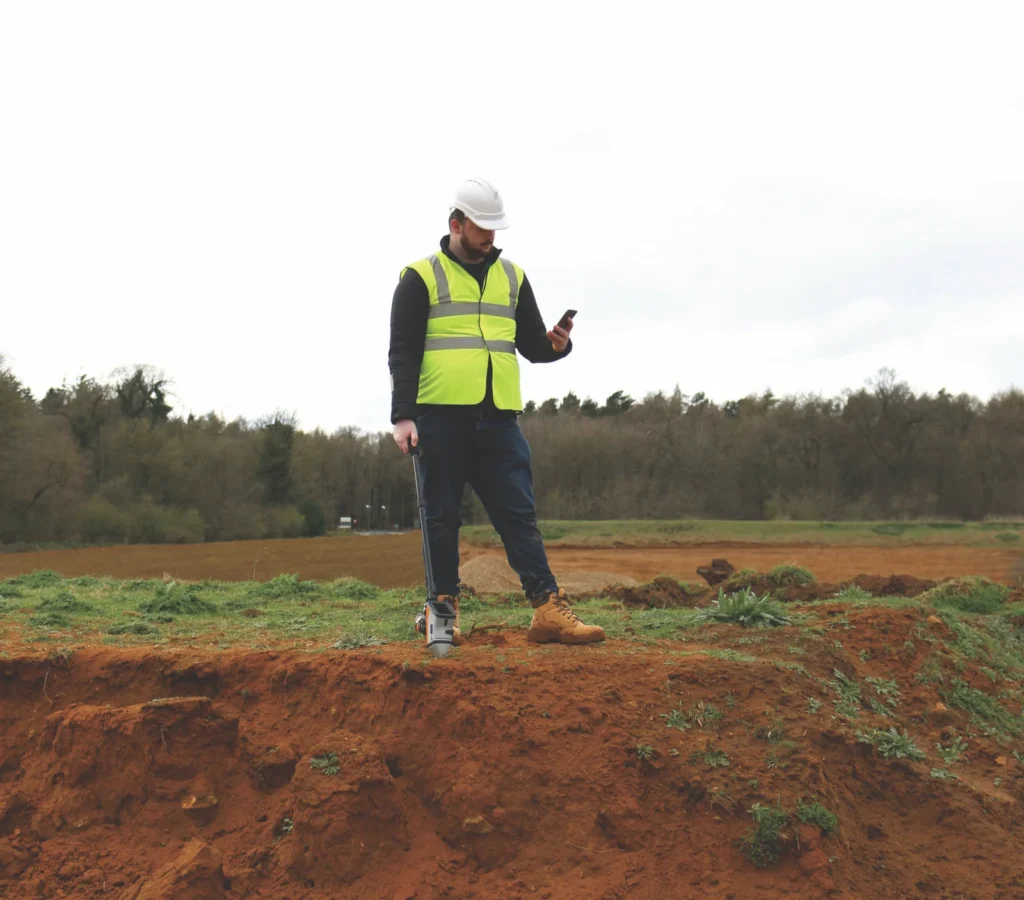 Un ingeniero utiliza un dispositivo portátil para el análisis de fluorescencia de rayos X en un entorno de construcción al aire libre, revisando datos en su teléfono móvil.