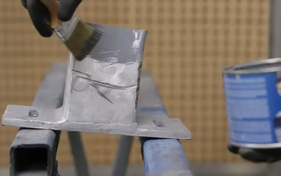 Una mano enguantada aplicando con brocha una capa de protección anticorrosiva gris sobre una pieza metálica, indicando el inicio del proceso de recubrimiento.