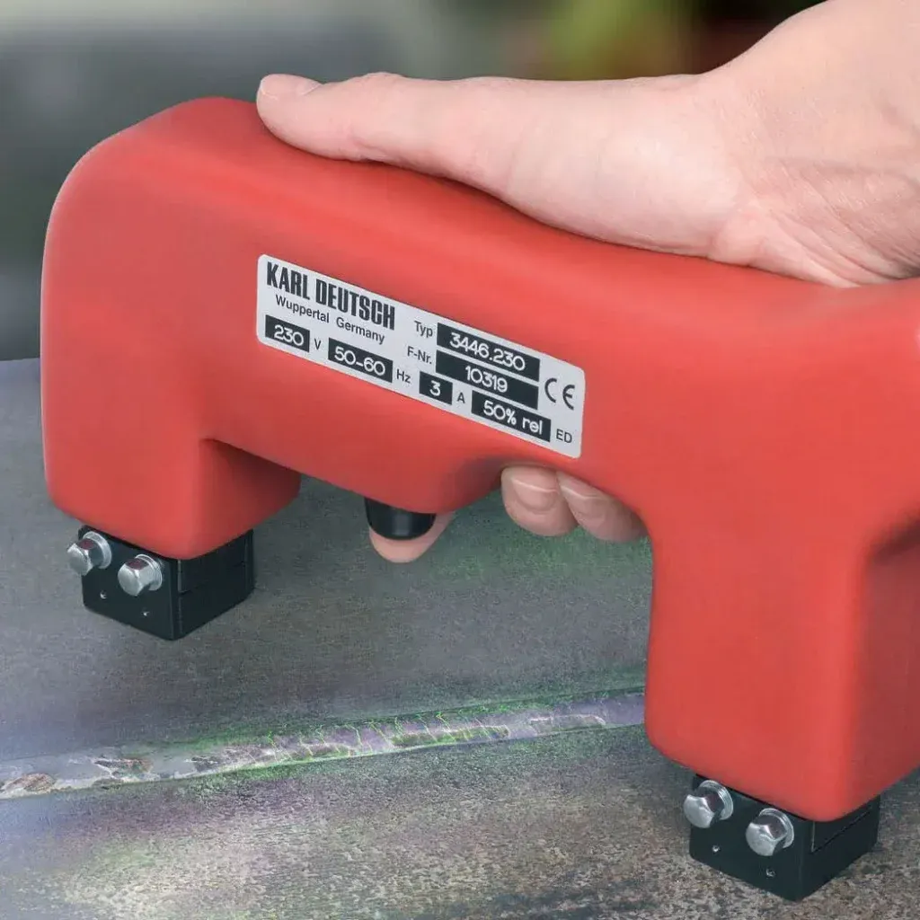 Una mano sostiene un yugo rojo de KARL DEUTSCH, herramienta para ensayo por partículas magnéticas, sobre una superficie metálica.