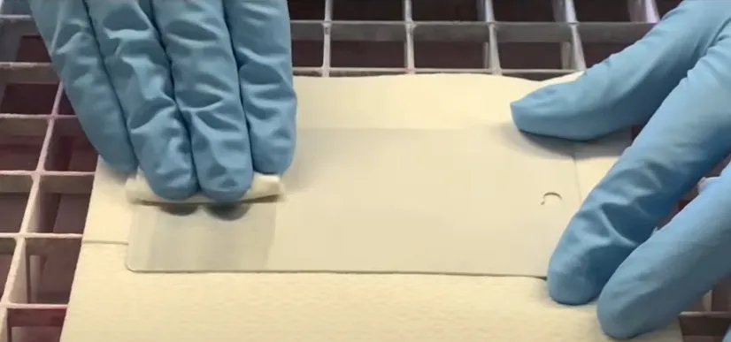 Manos enguantadas aplican cuidadosamente un desarrollador sobre una pieza metálica blanca, como parte de la inspección por líquidos penetrantes.