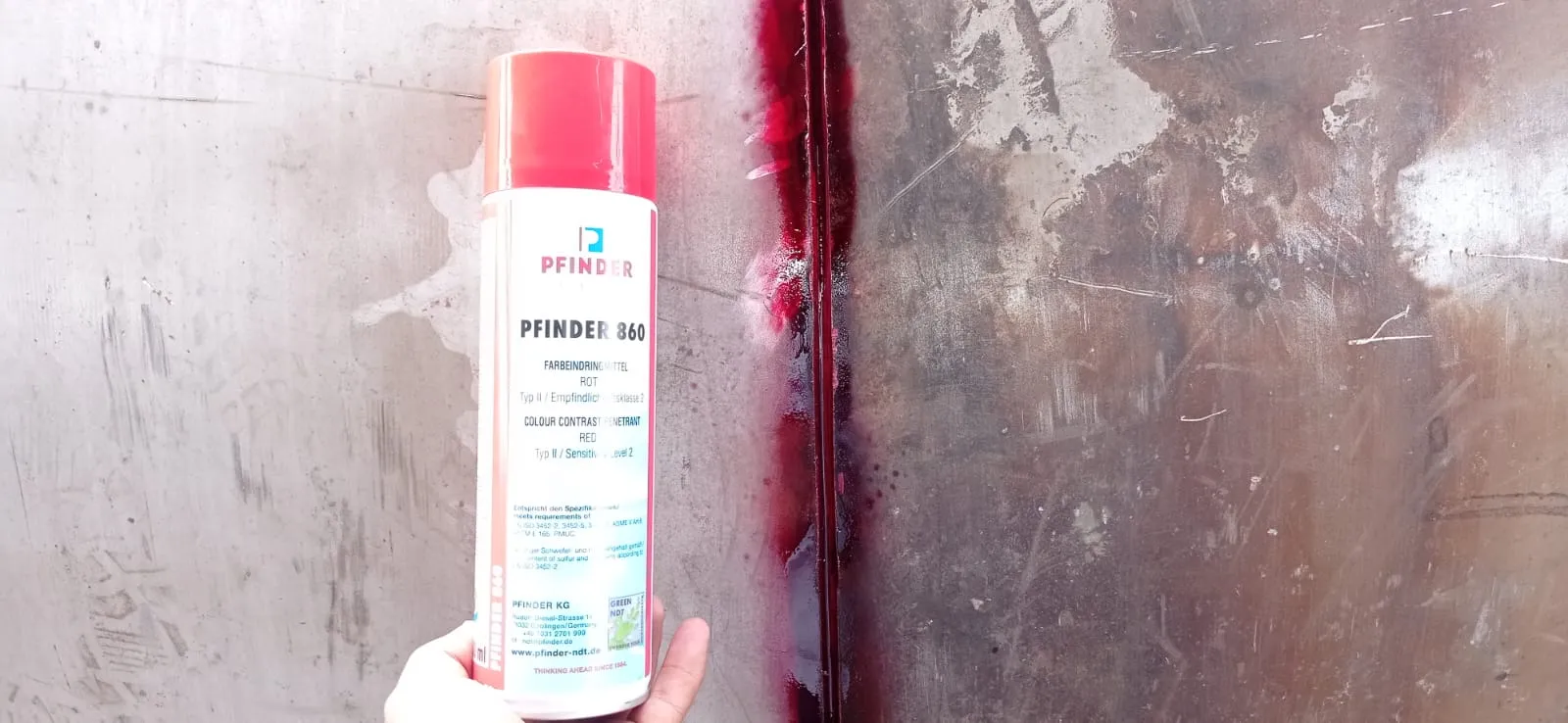 Una lata de aerosol de "PFINDER 860" líquido penetrante rojo se sostiene sobre una superficie metálica que muestra rastros del tinte rojo.