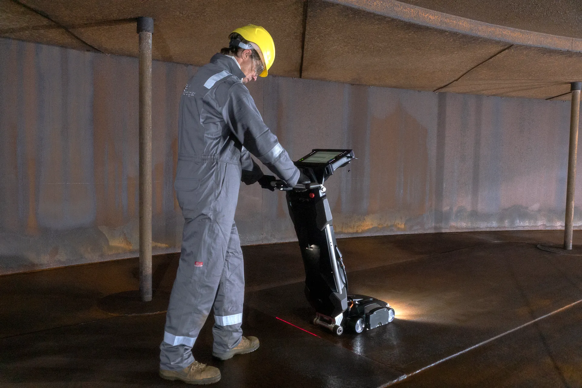Un operario con casco y overol utiliza un dispositivo de inspección por MFL portátil en una gran estructura metálica curvada.