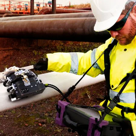 Un técnico de inspección con casco y chaleco de seguridad maneja un equipo de ensayo en una tubería, con otras tuberías industriales al fondo.