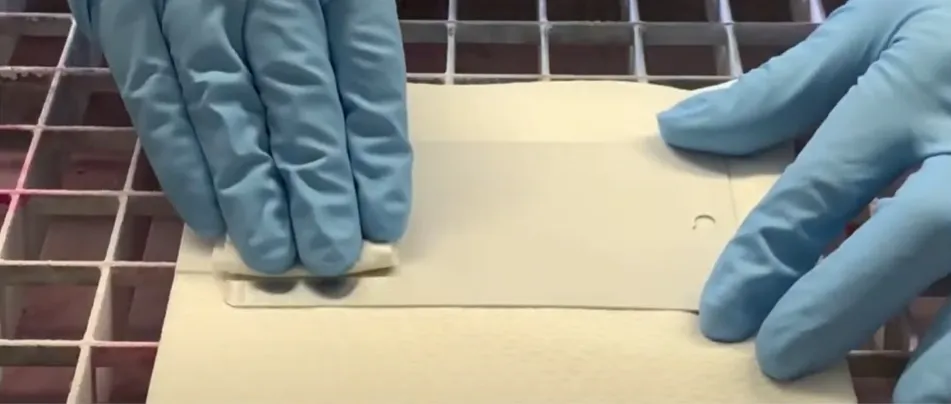 Una mano enguantada de azul desliza una pieza de cartón con revelador en polvo sobre una superficie metálica para esparcirlo uniformemente, revelando posibles indicaciones de defectos en el ensayo de tintas penetrantes.