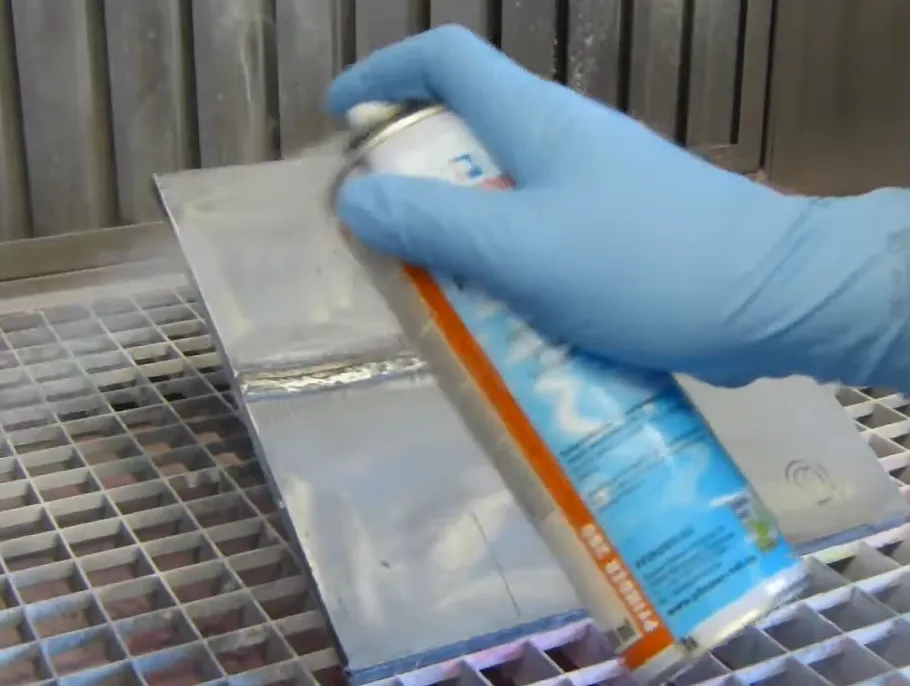 Una mano enguantada de azul sostiene una lata de spray y aplica un líquido sobre una superficie metálica plana que está sobre una rejilla, durante la fase inicial de un ensayo de tintas penetrantes.