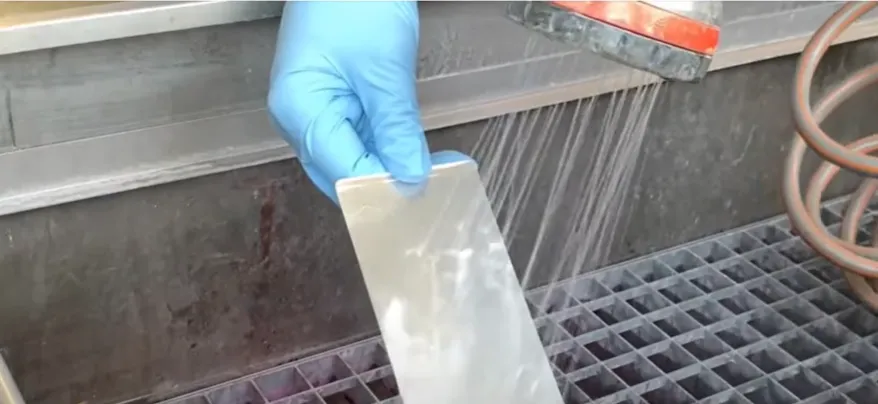 Una mano con un guante azul sostiene una pieza metálica plana bajo un chorro de agua, enjuagando el exceso de penetrante de la superficie durante un ensayo no destructivo.
