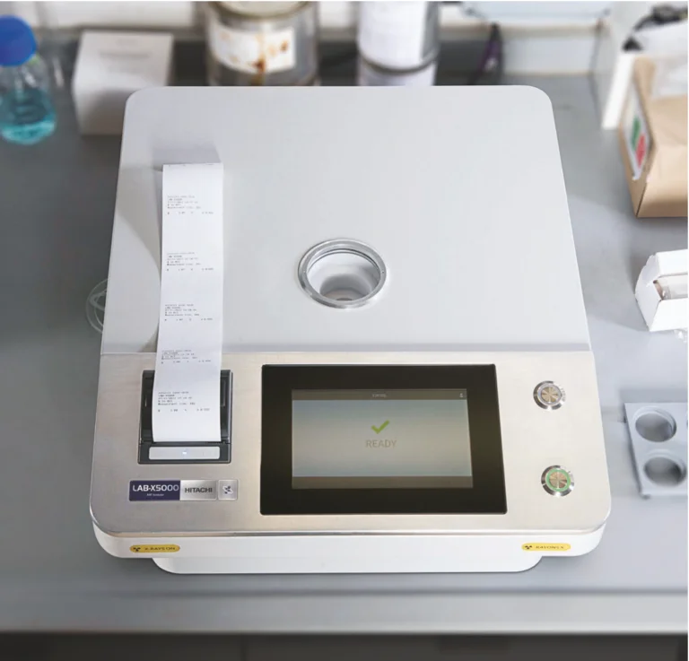 Primer plano de un analizador de fluorescencia de rayos X (XRF) de laboratorio con una pantalla "READY" e imprimiendo un ticket de resultados.