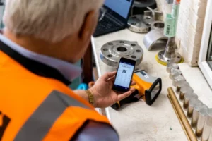 Vista trasera de un ingeniero de calidad consultando los resultados de un análisis de fluorescencia de rayos X (XRF) en su smartphone.