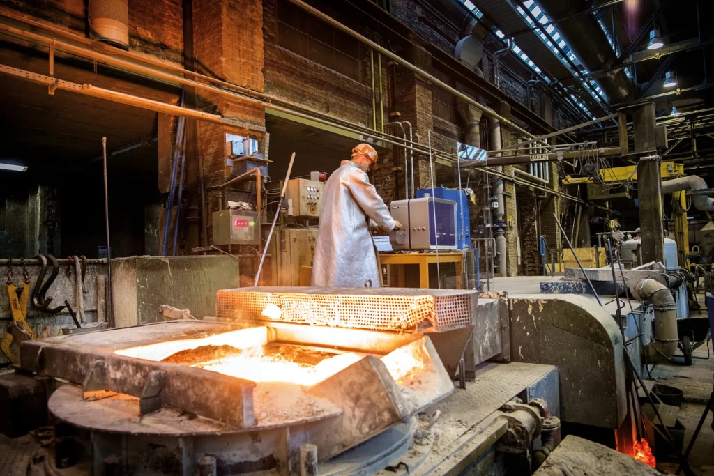 Un trabajador con equipo protector realiza tareas cerca de un horno de metal fundido en una fundición industrial.