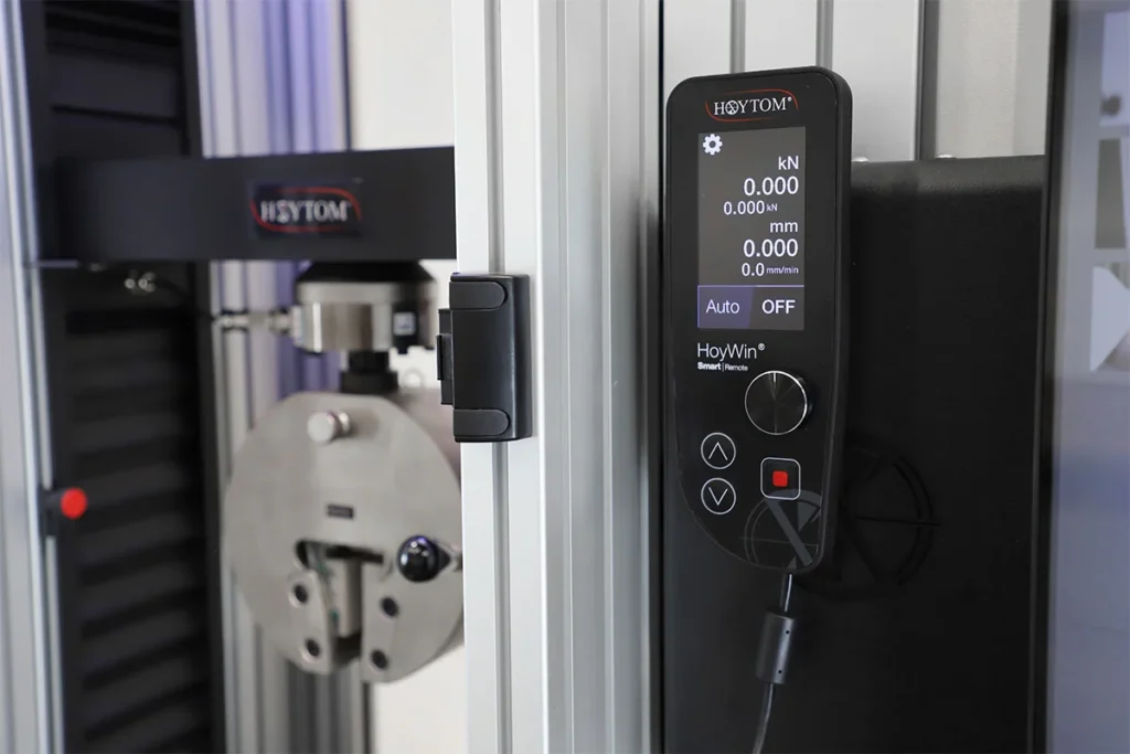 Detalle de la consola remota de control de la máquina de ensayo, mostrando en pantalla la carga, desplazamiento y velocidad del ensayo