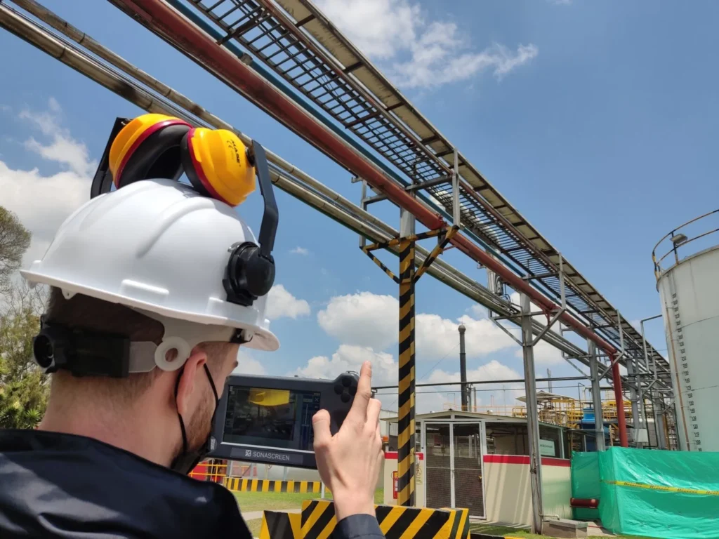 Un técnico de mantenimiento de equipo con casco y orejeras usa una tableta para inspeccionar tuberías industriales.