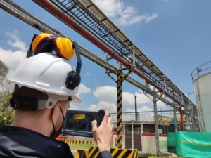 Un técnico de mantenimiento de equipo con casco y orejeras usa una tableta para inspeccionar tuberías industriales.