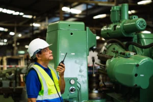Supervisor de fábrica usando radio cerca de maquinaria pesada bajo estrategias de integridad industrial.