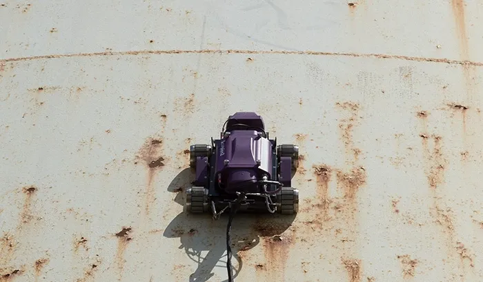 Robot oruga trepador realizando inspección automática con equipo de medición por ultrasonido industrial en tanque corroído.