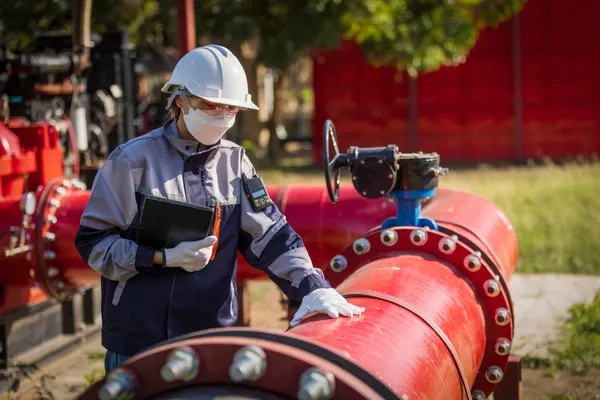 Inspectora con EPP y mascarilla revisa tubería roja de incendio con tableta, siguiendo protocolos API 570 en planta.