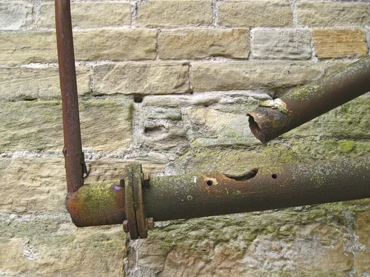 Tubo metálico roto y perforado mostrando los efectos severos de la corrosión por picaduras ante una pared de piedra.