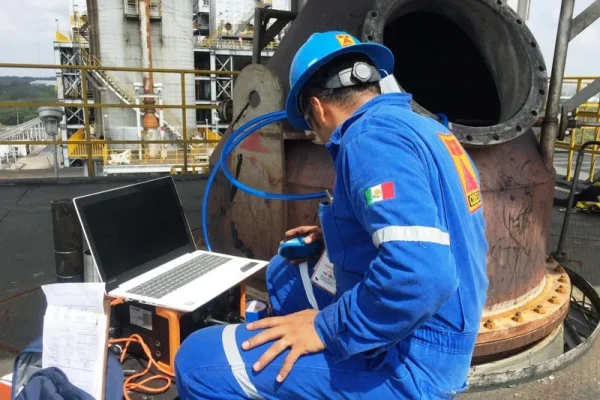 Operador de empresas de ensayos no destructivos en Perú analiza datos con laptop junto a un gran tanque industrial.