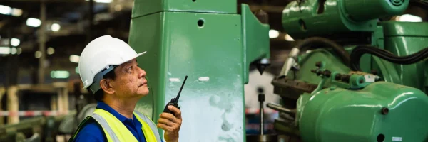 Supervisor de fábrica usando radio cerca de maquinaria pesada bajo estrategias de integridad industrial.